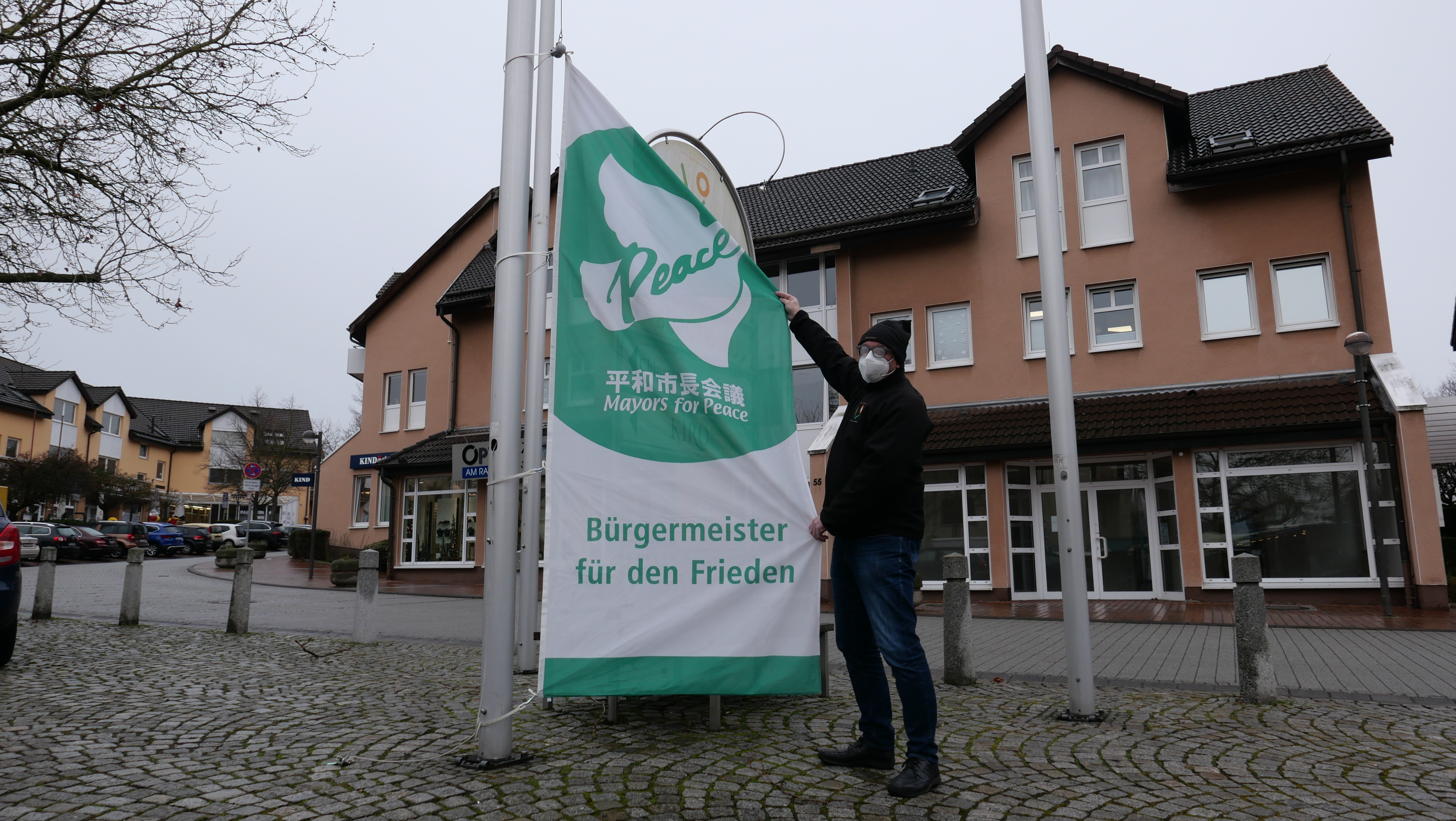 Auf dem Foto ist Bürgermeister der Gemeinde Lohfelden, Uwe Jäger, zu sehen, der die grün-weiße Mayors for Peace-Flagge auf dem Rathausplatz hisst. 