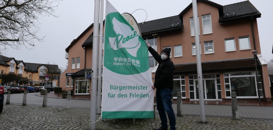 Auf dem Foto ist Bürgermeister der Gemeinde Lohfelden, Uwe Jäger, zu sehen, der die grün-weiße Mayors for Peace-Flagge auf dem Rathausplatz hisst. 