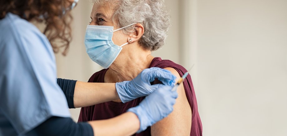 Auf dem Foto ist links eine junge Ärztin in einem hellblauen Kittel zu sehen, die einer Seniorin rechts neben in einem roten Pullover eine Corona-Schutzimpfung verabreicht.
