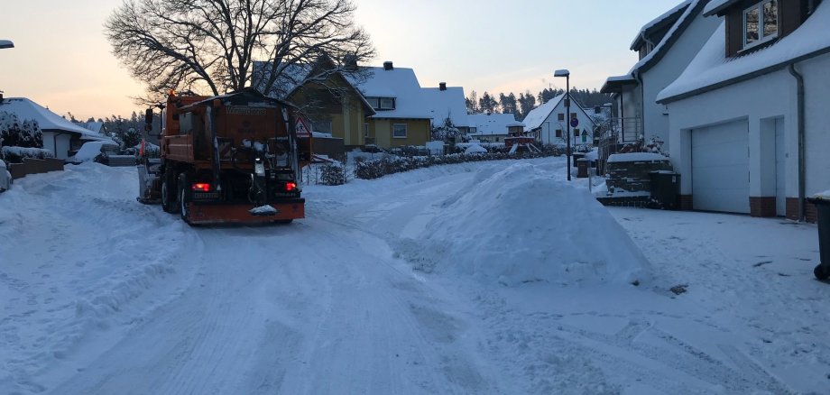 Auf dem Foto ist ein orangenes Winterdienst-Fahrzeug auf der linken Seite einer schneebedeckten Straßengabelung zu sehen. Auf der rechten Seite der Gabelung befindet sich rechts am Rand der Straße ein hoch aufgetürmter Schneeberg.