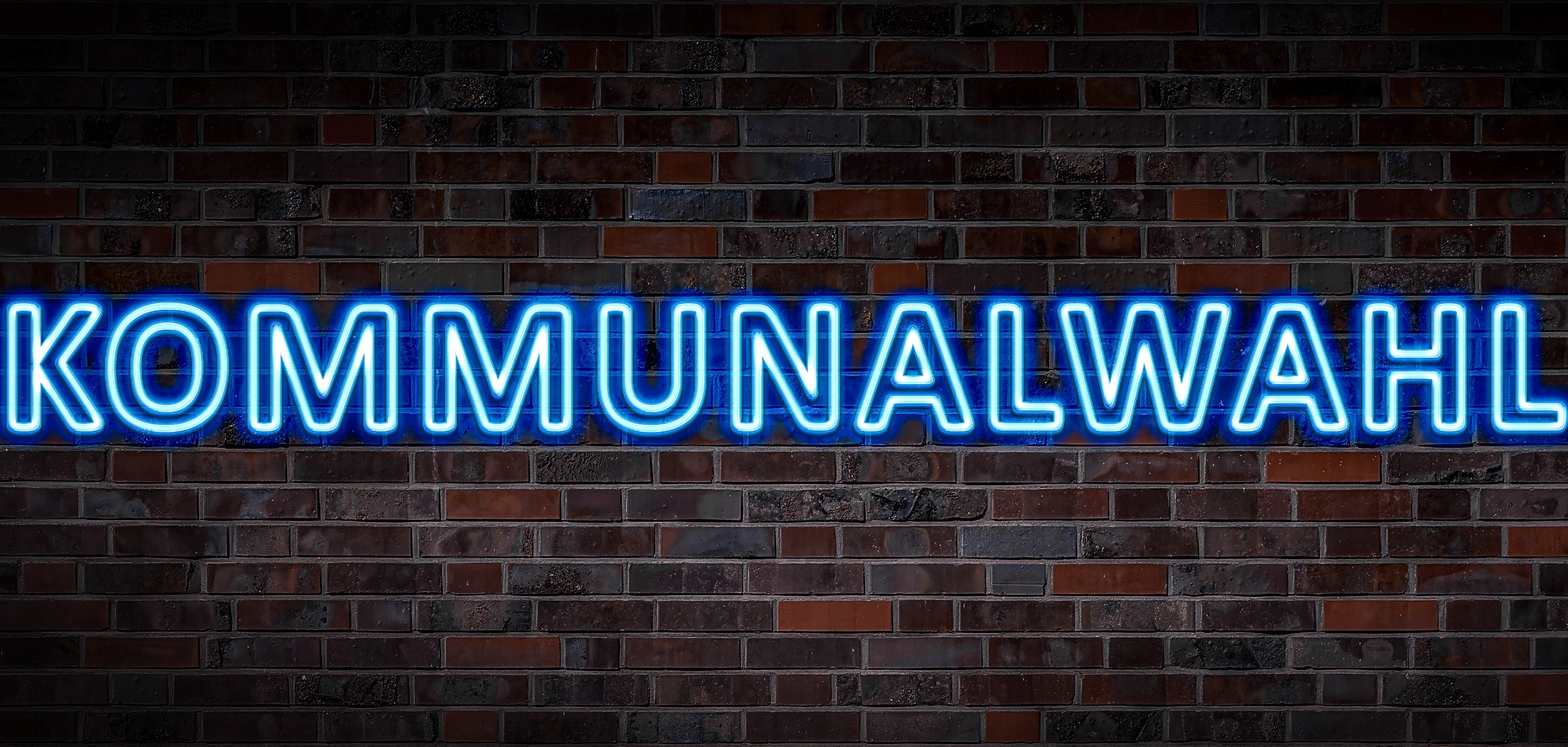 Auf dem Foto sieht man einen hellblauen Schriftzug mit der Aufschrift Kommunalwahlen auf einer dunkelbraunen Backsteinwand.