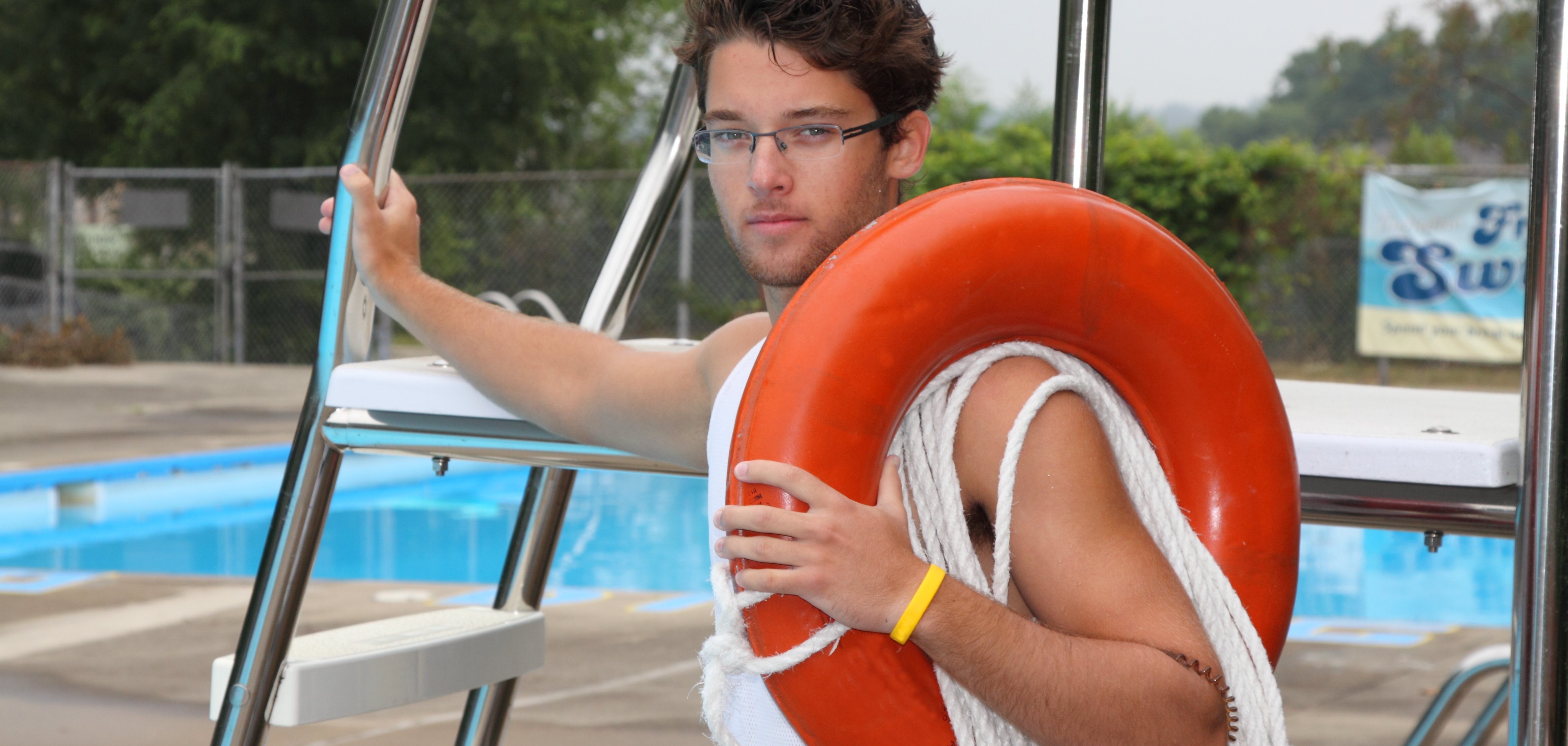 Auf dem Foto ist ein männlicher, junger Freibad-Mitarbeiter mit braunen Haaren und Brille zu sehen. Er trägt einen roten Rettungsreifen über dem linken Arm und ein gelbes Armband und steht vor einer Aussichtsplattform am Beckenrand eines Schwimmbeckens. Ein weiteres Schwimmbecken ist hinter dem ersten Becken zu erkennen. Im Hintergrund ist leicht verschwommen ein Schwimmbadzaun zu sehen, der teilweise von einer grünen Hecke, Bäumen und einem Werbebanner verdeckt wird.