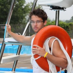Auf dem Foto ist ein männlicher, junger Freibad-Mitarbeiter mit braunen Haaren und Brille zu sehen. Er trägt einen roten Rettungsreifen über dem linken Arm und ein gelbes Armband und steht vor einer Aussichtsplattform am Beckenrand eines Schwimmbeckens. Ein weiteres Schwimmbecken ist hinter dem ersten Becken zu erkennen. Im Hintergrund ist leicht verschwommen ein Schwimmbadzaun zu sehen, der teilweise von einer grünen Hecke, Bäumen und einem Werbebanner verdeckt wird.
