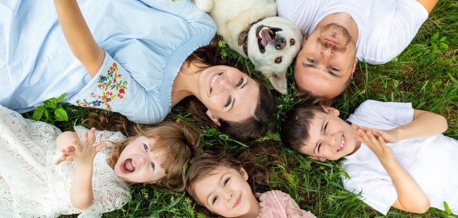 Eine junge, glückliche Familie, bestehend aus Vater, Mutter, zwei Mädchen, einem Jungen und einem weißen Hund, liegen auf dem Rasen und lächeln in die Kamera.