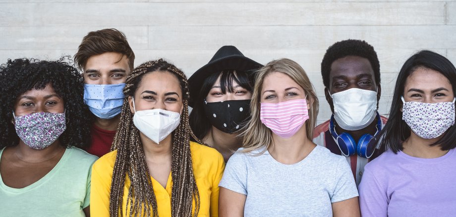 Auf dem Foto sind sieben Personen unterschiedlicher Nationalitäten zu sehen, die alle eine Corona-Schutzmaske tragen.