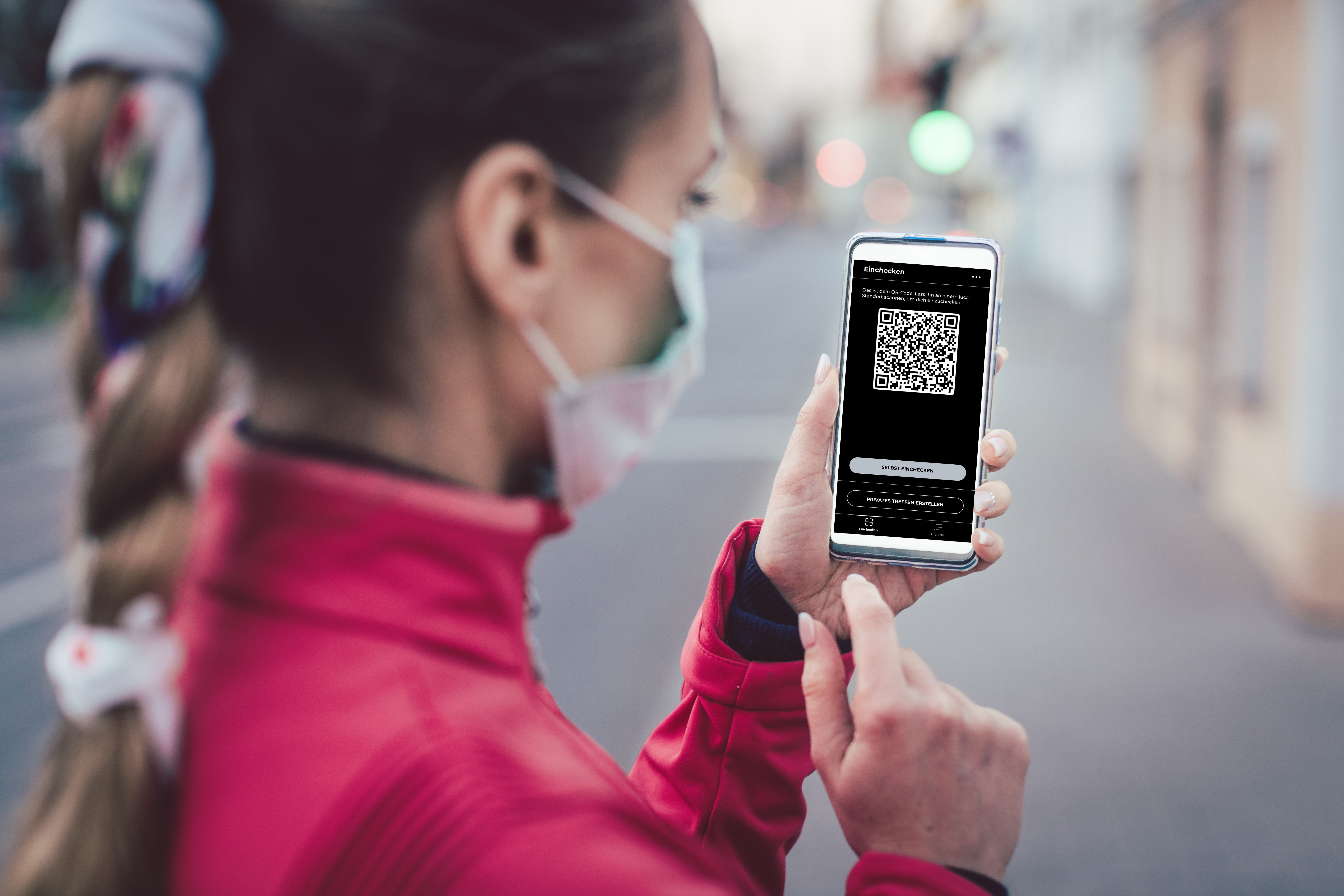 Auf dem Foto ist eine dunkelhaarige, junge Frau in einem pinken Rollkragenpullover zu sehen, die auf ihr Handy schaut, dessen Display die App luca anzeigt.