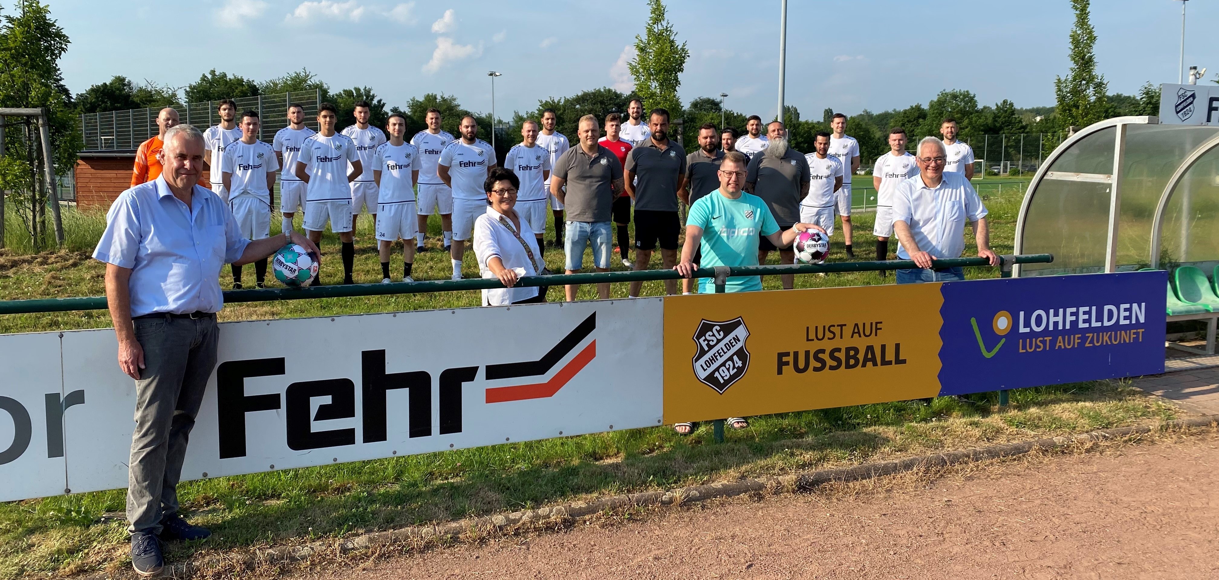 Die Spieler der 1. und 2. Mannschaft des FSC Lohfelden kurz vor ihrem ersten Training am 16.06.2021 mit FSC-Sponsoring-Beauftragter Herbert Brethauer, der Ersten Beigeordneten Bärbel Fehr, FSC-Hauptkassierer René Albert und Bürgermeister Uwe Jäger vor der neuen Bandenwerbung der Gemeinde Lohfelden im Nordhessen-Stadion.