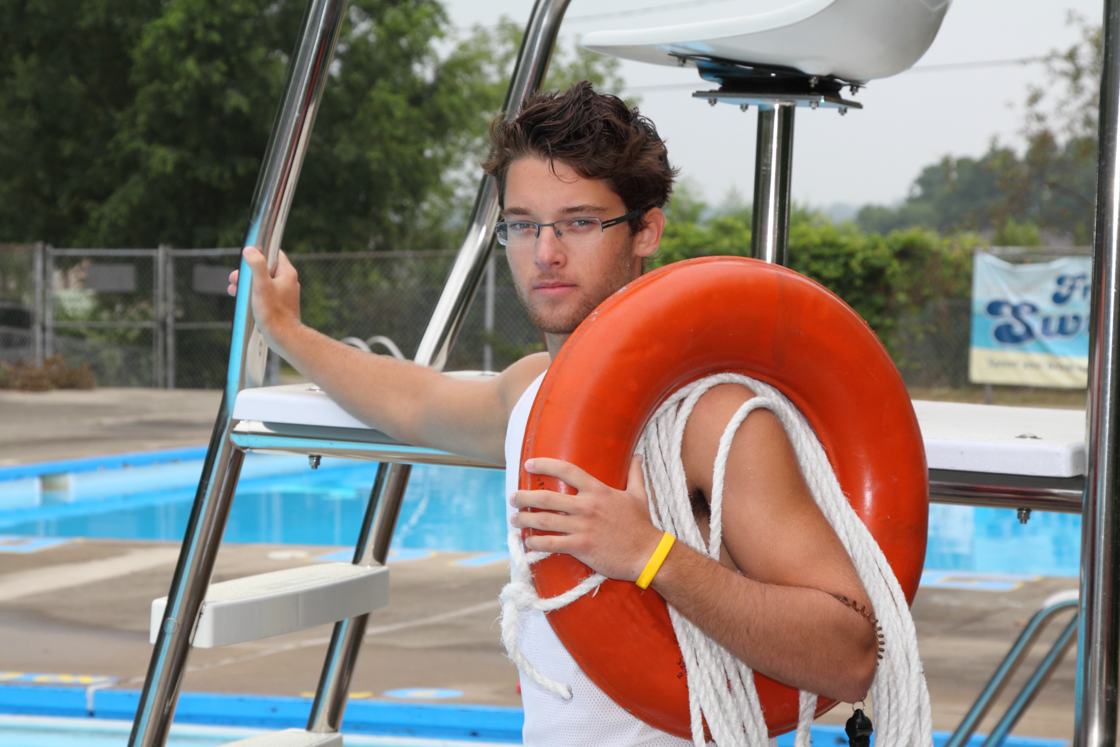 Auf dem Foto ist ein männlicher, junger Freibad-Mitarbeiter mit braunen Haaren und Brille zu sehen. Er trägt einen roten Rettungsreifen über dem linken Arm und ein gelbes Armband und steht vor einer Aussichtsplattform am Beckenrand eines Schwimmbeckens. Ein weiteres Schwimmbecken ist hinter dem ersten Becken zu erkennen. Im Hintergrund ist leicht verschwommen ein Schwimmbadzaun zu sehen, der teilweise von einer grünen Hecke, Bäumen und einem Werbebanner verdeckt wird.