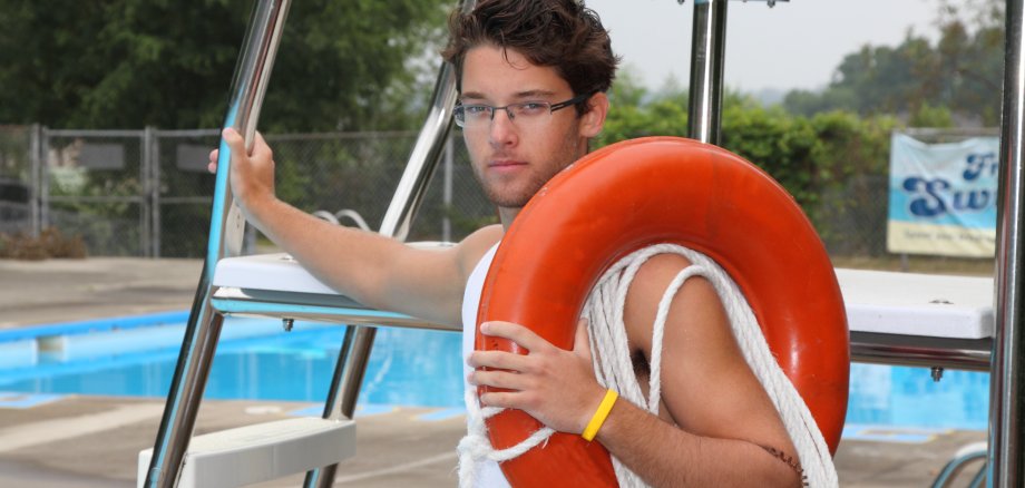 Auf dem Foto ist ein männlicher, junger Freibad-Mitarbeiter mit braunen Haaren und Brille zu sehen. Er trägt einen roten Rettungsreifen über dem linken Arm und ein gelbes Armband und steht vor einer Aussichtsplattform am Beckenrand eines Schwimmbeckens. Ein weiteres Schwimmbecken ist hinter dem ersten Becken zu erkennen. Im Hintergrund ist leicht verschwommen ein Schwimmbadzaun zu sehen, der teilweise von einer grünen Hecke, Bäumen und einem Werbebanner verdeckt wird.