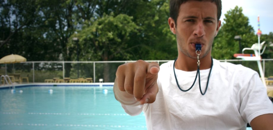 Auf dem Foto ist ein junger männlicher Freibad-Mitarbeiter zu sehen, der eine Trillerpfeife im Mund hat und einen Rettungsring im Arm hält. Im Hintergrund ist ein Schwimmbecken zu sehen. 