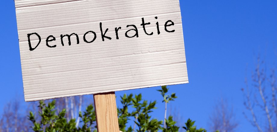 Auf dem Foto ist ein braunes Pappschild mit dem Schriftzug Demokratie zu lesen. Eine Hand hält den Holzgriff des Schildes in die Luft. Im Hintergrund sind ein blauer Himmel und die grünen Blätter eines Baumes zu sehen.