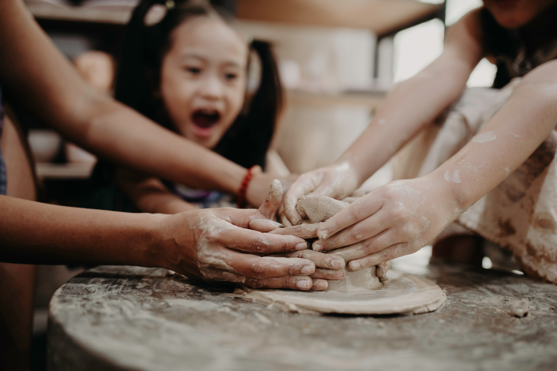 Auf dem Foto ist ein lachendes, dunkelhaariges Mädchen mit zwei Zöpfen zu sehen, die gemeinsam mit anderen Kindern und Erwachsenen töpfert.