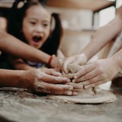 Auf dem Foto ist ein lachendes, dunkelhaariges Mädchen mit zwei Zöpfen zu sehen, die gemeinsam mit anderen Kindern und Erwachsenen töpfert.