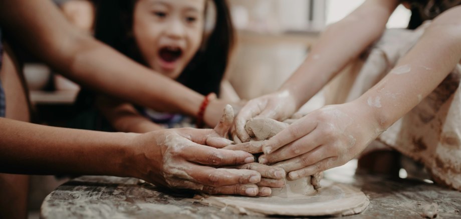 Auf dem Foto ist ein lachendes, dunkelhaariges Mädchen mit zwei Zöpfen zu sehen, die gemeinsam mit anderen Kindern und Erwachsenen töpfert.