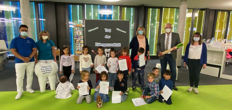 Zahnarzt Samiullah Habibi und seine Assistentin Gudrun Schmidt (links der Pinnwand) Bürgermeister Uwe Jäger und Büchereimitarbeiterinnen Gabriele Nemenz und Michaela Fenn (rechts der Pinnwand), mit den Kindern der Kindertagesstätte des ASB beim „Tag der Zahngesundheit“ in der Gemeinde- und Schulbücherei Lohfelden.
