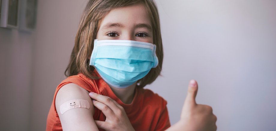Auf dem Foto ist ein lächelndes kleines Kind mit einer medizinischen Gesichtsmaske kurz nach seiner Covid-19-Impfung zu sehen, das ein kleines Pflaster mit einem Lachsmiley auf dem rechten Arm trägt und den linken Daumen zu einem Okay in die Lust streckt.