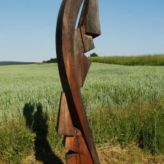 Die Skulptur "Gemeinsame Wege" vom Berger Künstler Werner Pirker ist aus Holz und zeigt zwei abstrakte Gesichter die gemeinsam zurück und nach vorn sehen.  Das Kunstwerk steht am ARS Natura x3 oberhalb der Wanderparkplatzes Herchenbach.