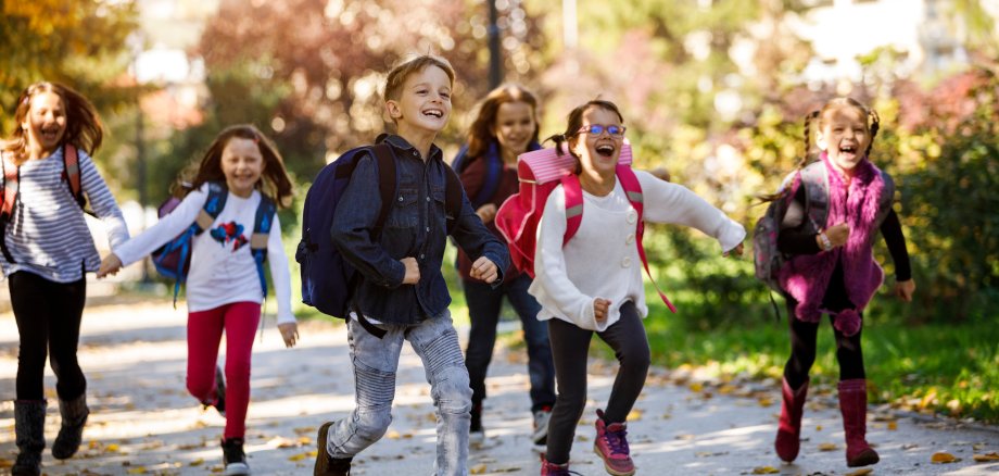 Auf dem Foto ist eine Gruppe von jungen Schulkindern zu sehen, die fröhlich auf einem Weg entlanglaufen und Schulranzen tragen.