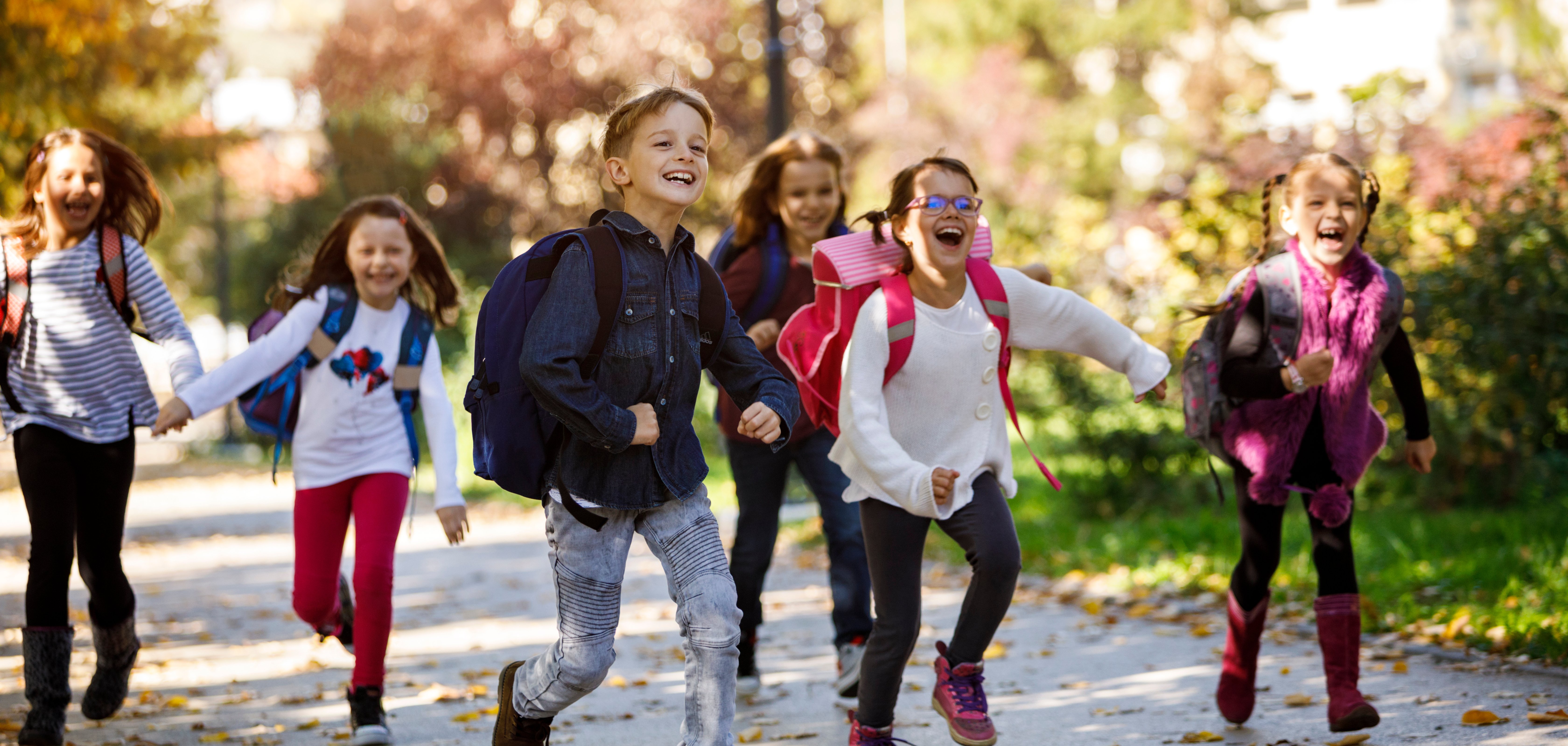 Auf dem Foto ist eine Gruppe von jungen Schulkindern zu sehen, die fröhlich auf einem Weg entlanglaufen und Schulranzen tragen.