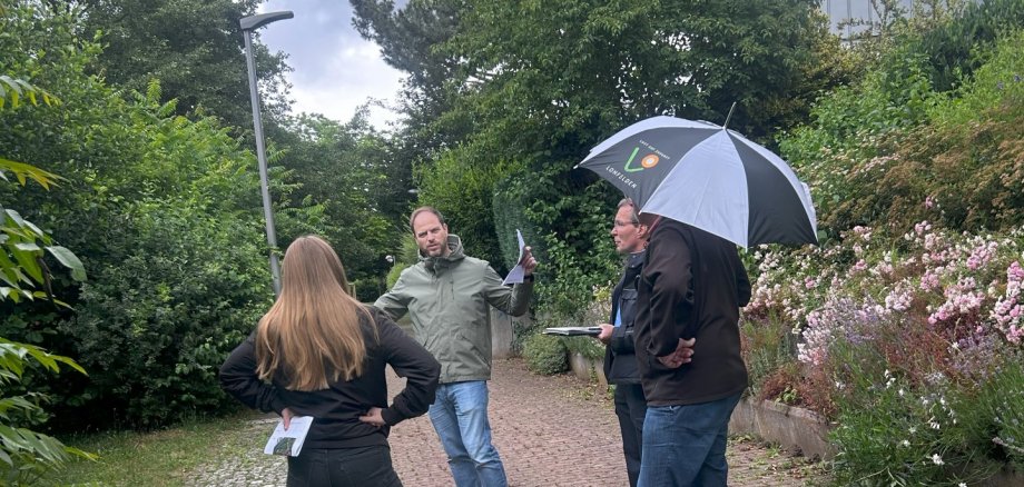 Eine Gruppe bestehend aus einer weblichen und drei männlichen Personen mittleren Alters stehen nebeneinander bei durchwachsenem Wetter auf dem Gehweg an einem See; Die mittlere Person erklärt den anderen Personen etwas.