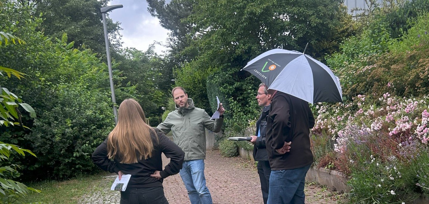 KOMPASS-Berater des Polizeipräsidiums Nordhessen, Kriminalhauptkommissar Mirco Skrzipek (M.), gemeinsam mit einem Teil der Arbeitsgruppe während der Ortsbegehung am 8. Juli im Rahmen der KOMPASS-Initiative. Eine Gruppe bestehend aus einer weblichen und drei männlichen Personen mittleren Alters stehen nebeneinander bei durchwachsenem Wetter auf dem Gehweg an einem See; Die mittlere Person erklärt den anderen Personen etwas.