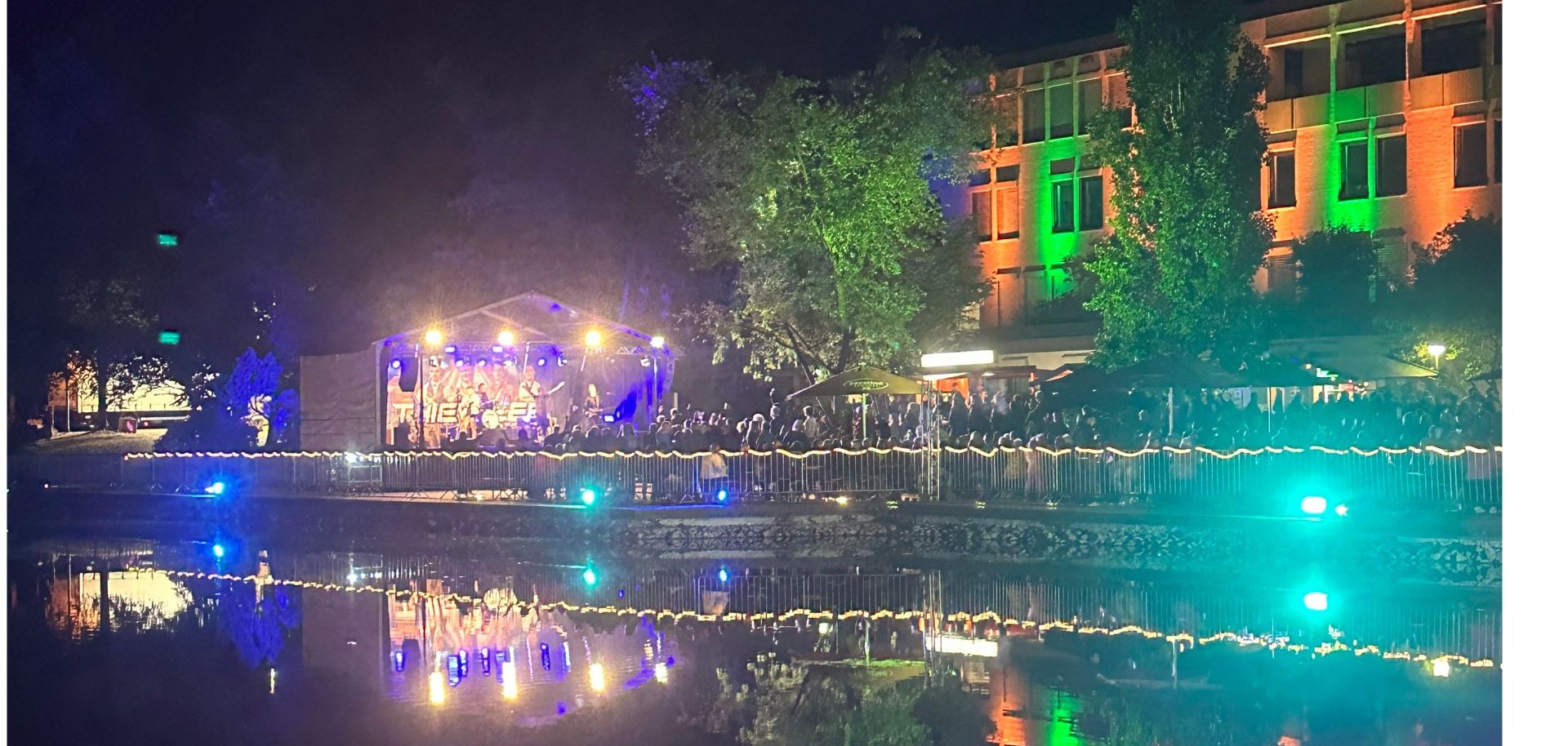 Rathaus Lohfelden im Dunklen, angestrahlt in orange und grün; Wasser im Vordergrund; beleuchtete Bühne links.