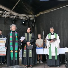 2 Pfarrerinnen und 1 Pfarrer in Talaren auf der Bühne.