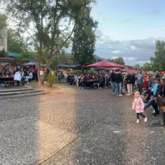 Menschen an Bierzeltgarnituren am befestigten Seeufer.