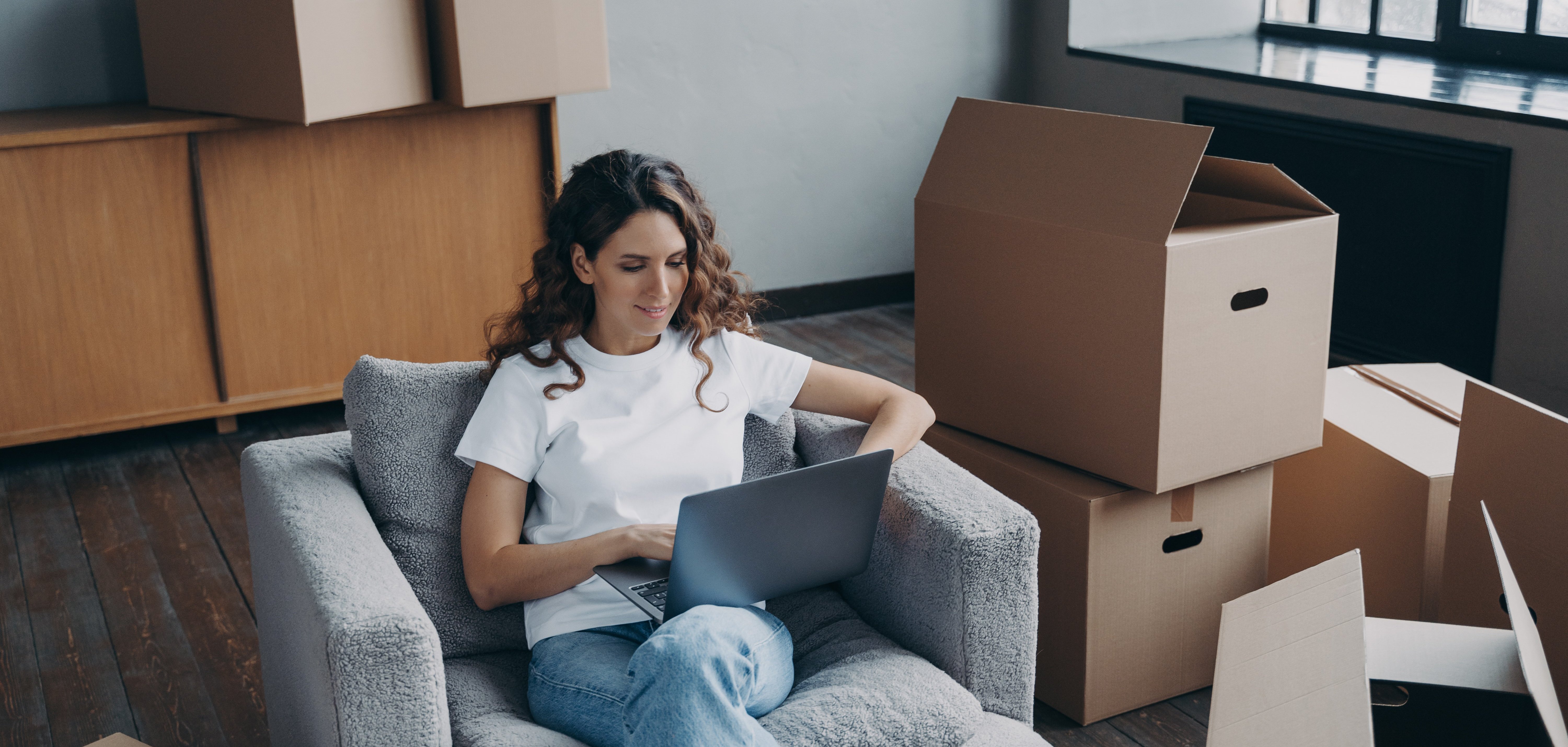 Glückliche junge Frau meldet sich in ihrer neuen Wohnung online um. Sie sitzt mit Laptop zwischen Kartons und lächelt.