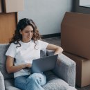 Glückliche junge Frau meldet sich in ihrer neuen Wohnung online um. Sie sitzt mit Laptop zwischen Kartons und lächelt.