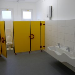 Container-Anlage - kleine Toiletten, gelbe Abtrennungen mit blauen Griffen, im Vordergrund kleine weiße Waschbecken in zwei verschiedenen Höhen.