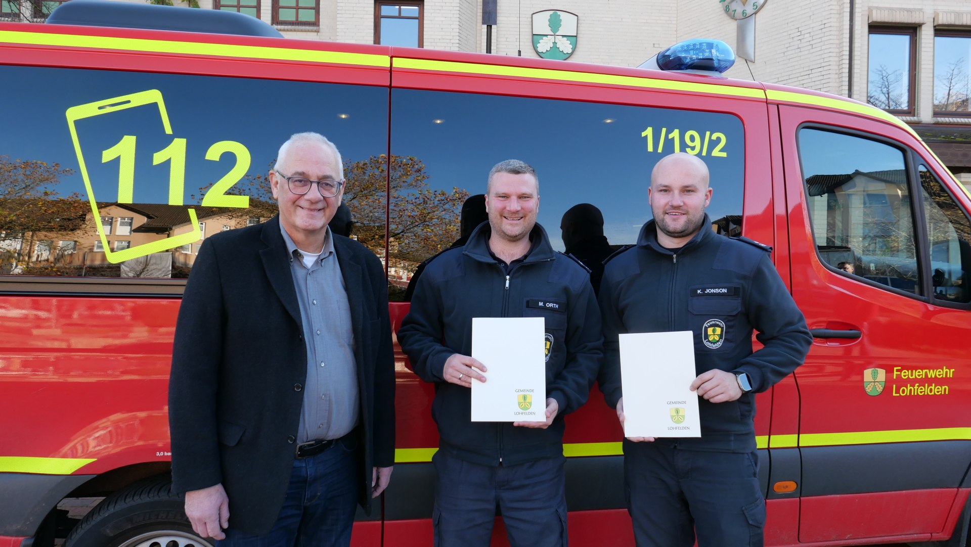 Zwei junge Männer in Feuerwehrkleidung stehen gemeinsam mit einem Mann mittleren Alters im Anzug vor einem roten Feuerwehrfahrzeug; in ihren Händen halten die beiden jungen Männer ihre Ernennungsurkunden.