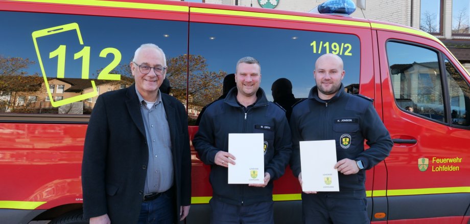Zwei junge Männer in Feuerwehrkleidung stehen gemeinsam mit einem Mann mittleren Alters im Anzug vor einem roten Feuerwehrfahrzeug; in ihren Händen halten die beiden jungen Männer ihre Ernennungsurkunden.