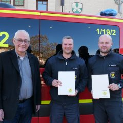 Zwei junge Männer in Feuerwehrkleidung stehen gemeinsam mit einem Mann mittleren Alters im Anzug vor einem roten Feuerwehrfahrzeug; in ihren Händen halten die beiden jungen Männer ihre Ernennungsurkunden.