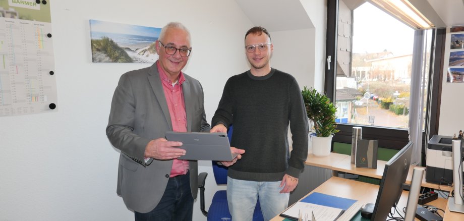 Ein junger Auszubildender in grauer Jeans und schwarzem Pullover steht neben einem Mann mittleren Alters im grauen Jacket und rosanen Hemd und mit einem Tablet in der Hand vor einem Schreibtisch in einem Büro einer Gemeindeverwaltung.