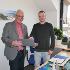 Ein junger Auszubildender in grauer Jeans und schwarzem Pullover steht neben einem Mann mittleren Alters im grauen Jacket und rosanen Hemd und mit einem Tablet in der Hand vor einem Schreibtisch in einem Büro einer Gemeindeverwaltung.