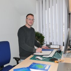 Ein junger Auszubildender in grauer Jeans und schwarzem Pullover steht vor seinem Schreibtisch in einem Büro einer Gemeindeverwaltung; er hält einen Stempel in der Hand mit dem er einen Becheid abstempelt.