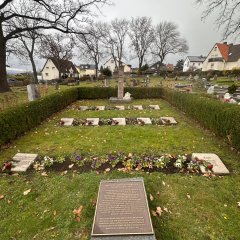 Grüber mit Grabsteinen darüber, umfasst mit einer kleinen Hecke. Im Fordergrund der Gedenkstein mit der Beschreibung des Unglücks.