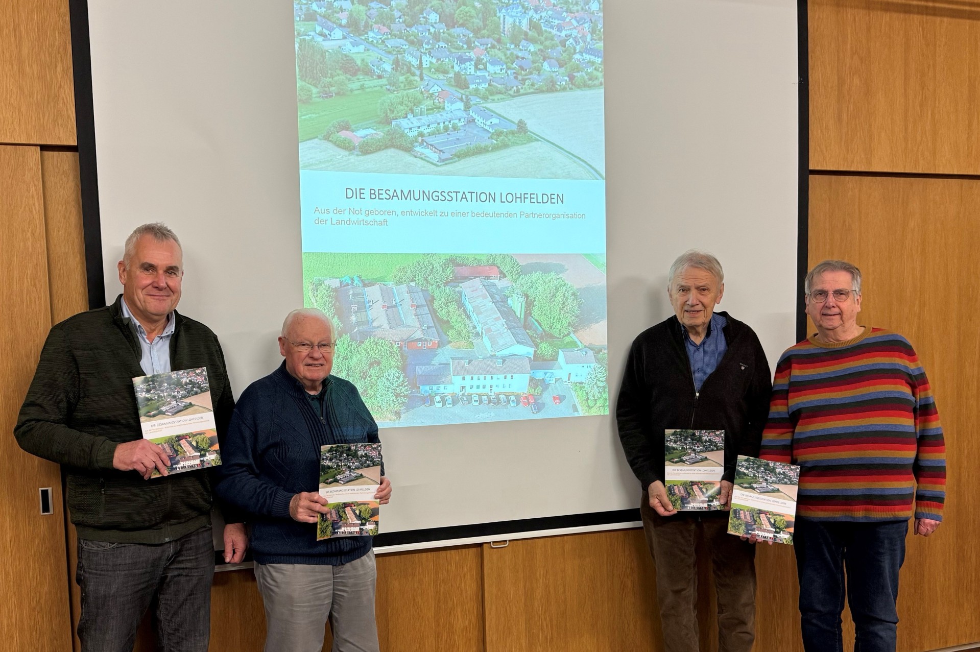Vier Männer höheren Alters in Freizeitkleidung stehen vor einer Beamer-Leinwand auf der das Foto der Besamungsstation Lohfelden projiziert ist; jeder der vier Herren hält ein Buch über die Besamungsstation Lohfelden in der Hand.