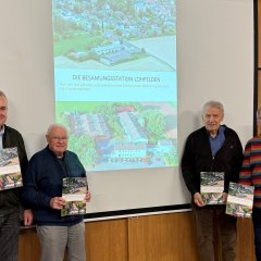 Vier Männer höheren Alters in Freizeitkleidung stehen vor einer Beamer-Leinwand auf der das Foto der Besamungsstation Lohfelden projiziert ist; jeder der vier Herren hält ein Buch über die Besamungsstation Lohfelden in der Hand.