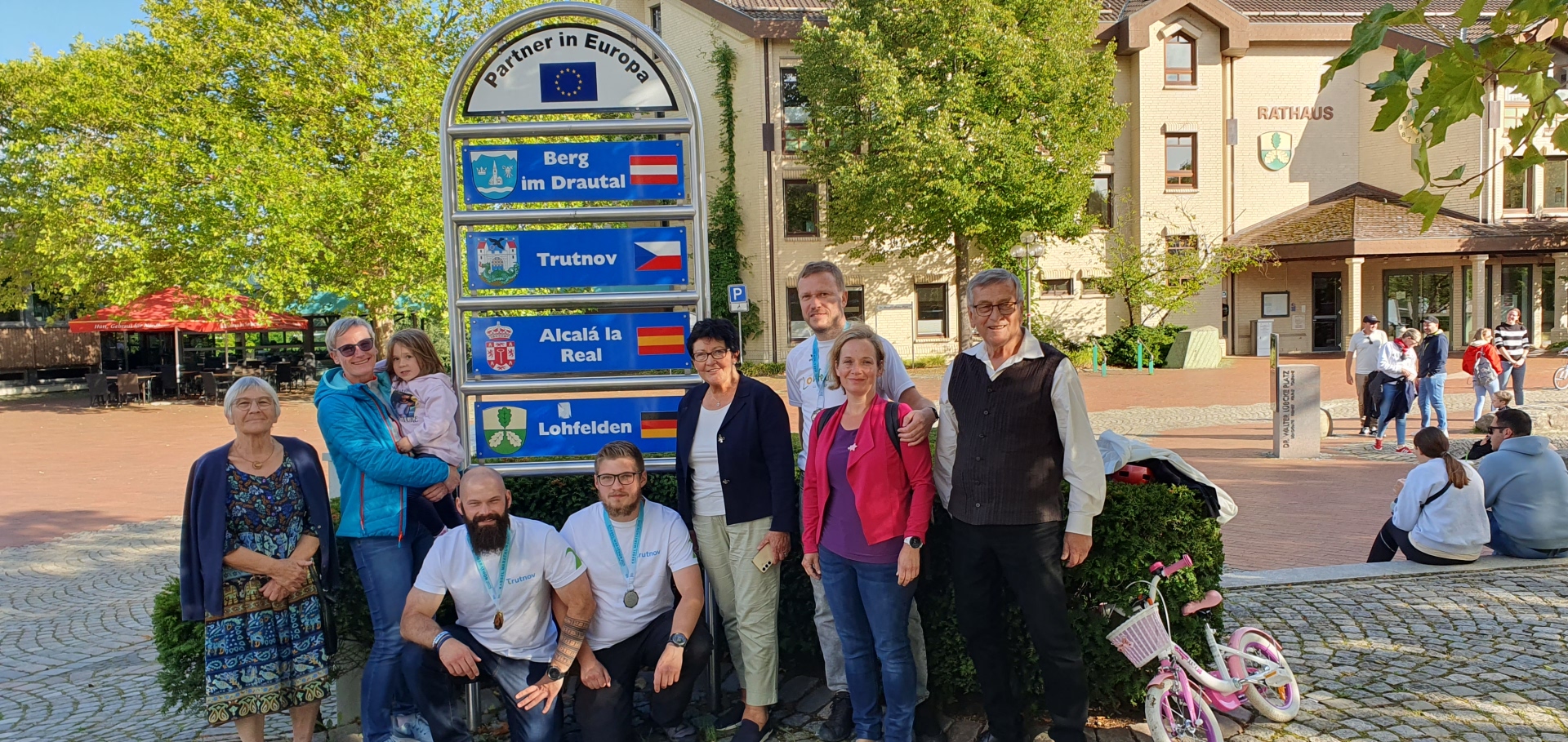 Nach dem Marathon fanden ein Rundgang durch Lohfelden sowie ein Abschiedsessen im Restaurant Vietnam Plus statt (v.l.n.r.): Agenda-Mitglied Marie-José Kuhn, Agenda-Sprecherin Eva Eichholz mit Natalie Fryč, die tschechischen Läufer Vojtěch Hubka und Vít Hubka, Erste Beigeordnete Bärbel Fehr, Läufer und Agenda-Mitglied und Läufer David Fryč, Agenda-Mitglied Vaclava Fryč und Agenda-Mitglied Kalle Kuhn. Eine Gruppe bestehend aus neun Männern und Frauen verschiedener Altersgruppen sowie einem Kind steht an einem sonnigen Tag nebeneinander auf dem Vorplatz eines Rathauses.