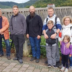 Eine Gruppe bestehend aus sechs Personen mittleren Alters sowie zwei Kindern posiert bei bewölktem Wetter nebeneinander vor der Sperrmauer eines Stausees.