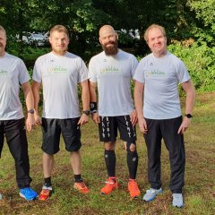 Eine Gruppe bestehend aus vier männlichen Marathon-Läufern jüngeren Alters steht nebeneinander in weißen Laufshirts an einem sonnigen Tag auf einer Wiese; Im Hintergrund stehen Bäume.