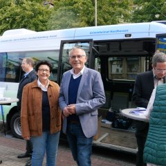 Eine Gruppe von Menschen stehen locker verteilt vor einem Bürgerbus an einer Einkaufsstraße; im Vordergrund deutlich zu erkennen eine Frau und ein Mann mittleren Alters.