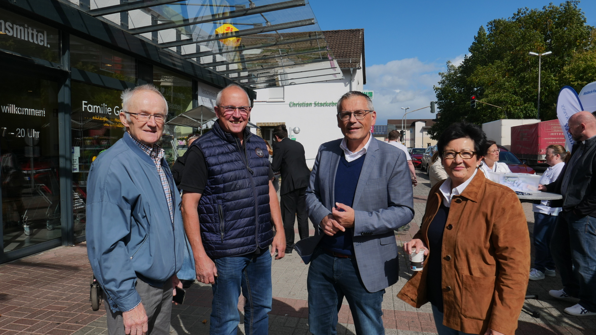 Eine Gruppe bestehend aus einer Frau und drei Männern mittleren Alters stehen bei Sonnenschein nebeneinander vor einem Lebensmittelgeschäft an einer Einkaufsstraße.