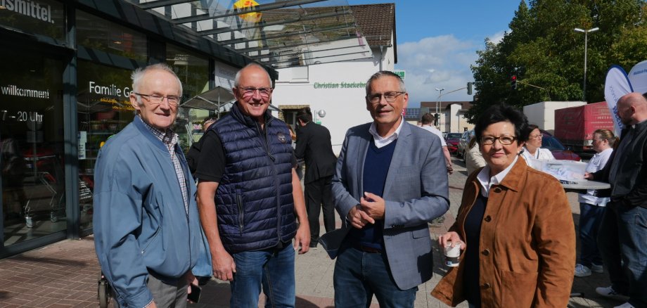 Eine Gruppe bestehend aus einer Frau und drei Männern mittleren Alters stehen bei Sonnenschein nebeneinander vor einem Lebensmittelgeschäft an einer Einkaufsstraße.
