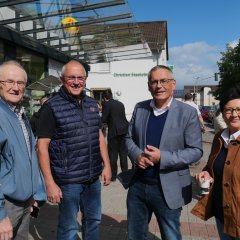 Eine Gruppe bestehend aus einer Frau und drei Männern mittleren Alters stehen bei Sonnenschein nebeneinander vor einem Lebensmittelgeschäft an einer Einkaufsstraße.