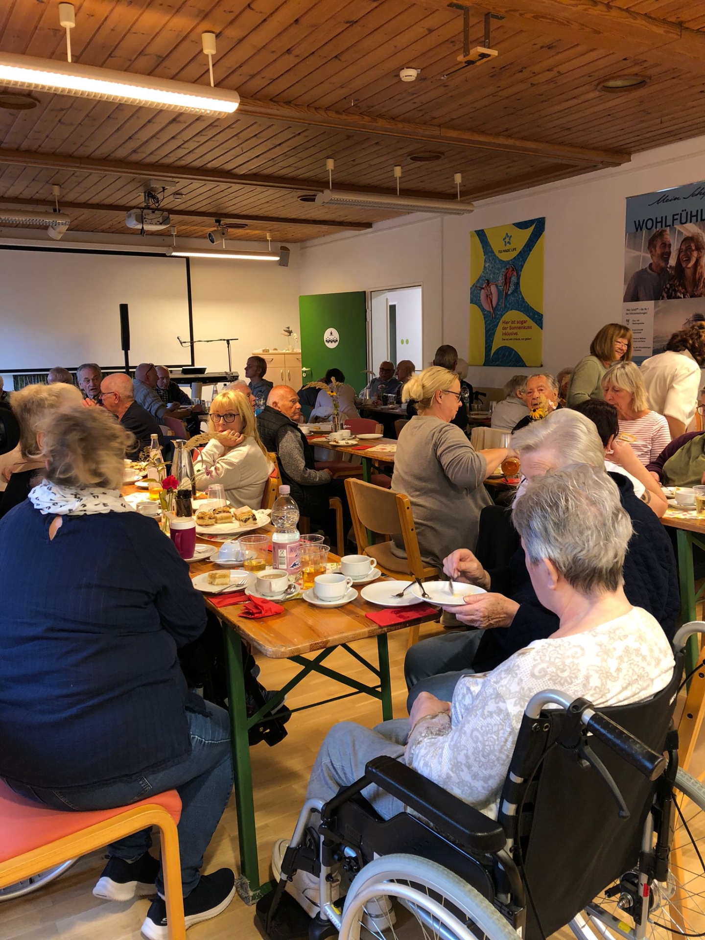 Eine größere Gruppe von Seniorinnen und Senioren sitzt beim gemeinsamen Kaffeetrinken an langen Tischen im Mehrzweckraum eines Seniorenwohnheims.