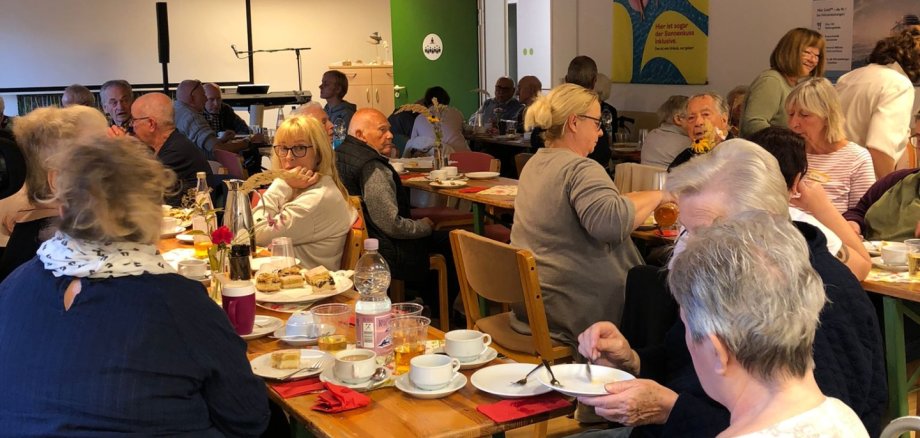 Eine größere Gruppe von Seniorinnen und Senioren sitzt beim gemeinsamen Kaffeetrinken an langen Tischen im Mehrzweckraum eines Seniorenwohnheims.