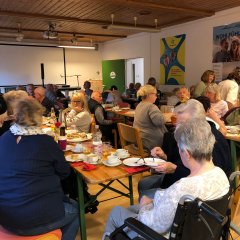 Eine größere Gruppe von Seniorinnen und Senioren sitzt beim gemeinsamen Kaffeetrinken an langen Tischen im Mehrzweckraum eines Seniorenwohnheims.